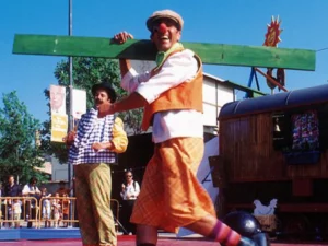Imagen de dos payasos llenan de alegría y risas una actuación al aire libre. Uno de ellos, con una gran sonrisa y un colorido atuendo, carga una tabla verde sobre sus hombros mientras su compañero, también vestido de manera extravagante, observa con diversión. El ambiente está impregnado de entusiasmo y emoción, con el público disfrutando del espectáculo en un día soleado. Estas actuaciones no solo entretienen, sino que también inspiran y despiertan la creatividad de los más pequeños, dejando una huella imborrable en su memoria. Correspondiente a la categoría de talleres creativos y experiencias para regalar a niños en Barcelona - Regalexia.
