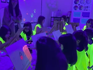 En la imagen, se observa a un grupo de niñas concentradas en su labor artística, pintando con colores neón bajo la luz ultravioleta. Las niñas, vestidas con chalecos de seguridad fluorescentes, están sentadas en pequeños caballetes, cada una con su propia obra en progreso. Correspondiente a la categoría de tallesres creativos. -Sitios para celebrar fiestas con niños en Barcelona
